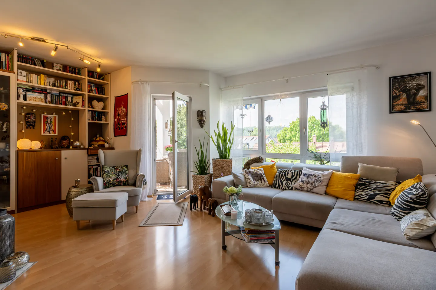 Bright living room with wood floors, a beige sectional sofa with patterned pillows, and a bookshelf filled with books and decorative items. Balcony access.