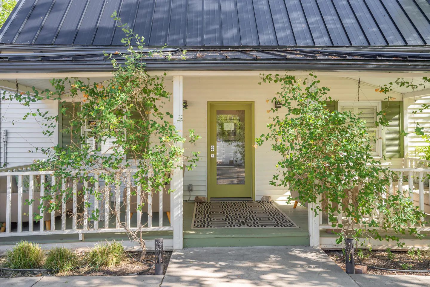 Front view of a white house with a green door and shutters, a black metal roof, and climbing plants on the porch.
