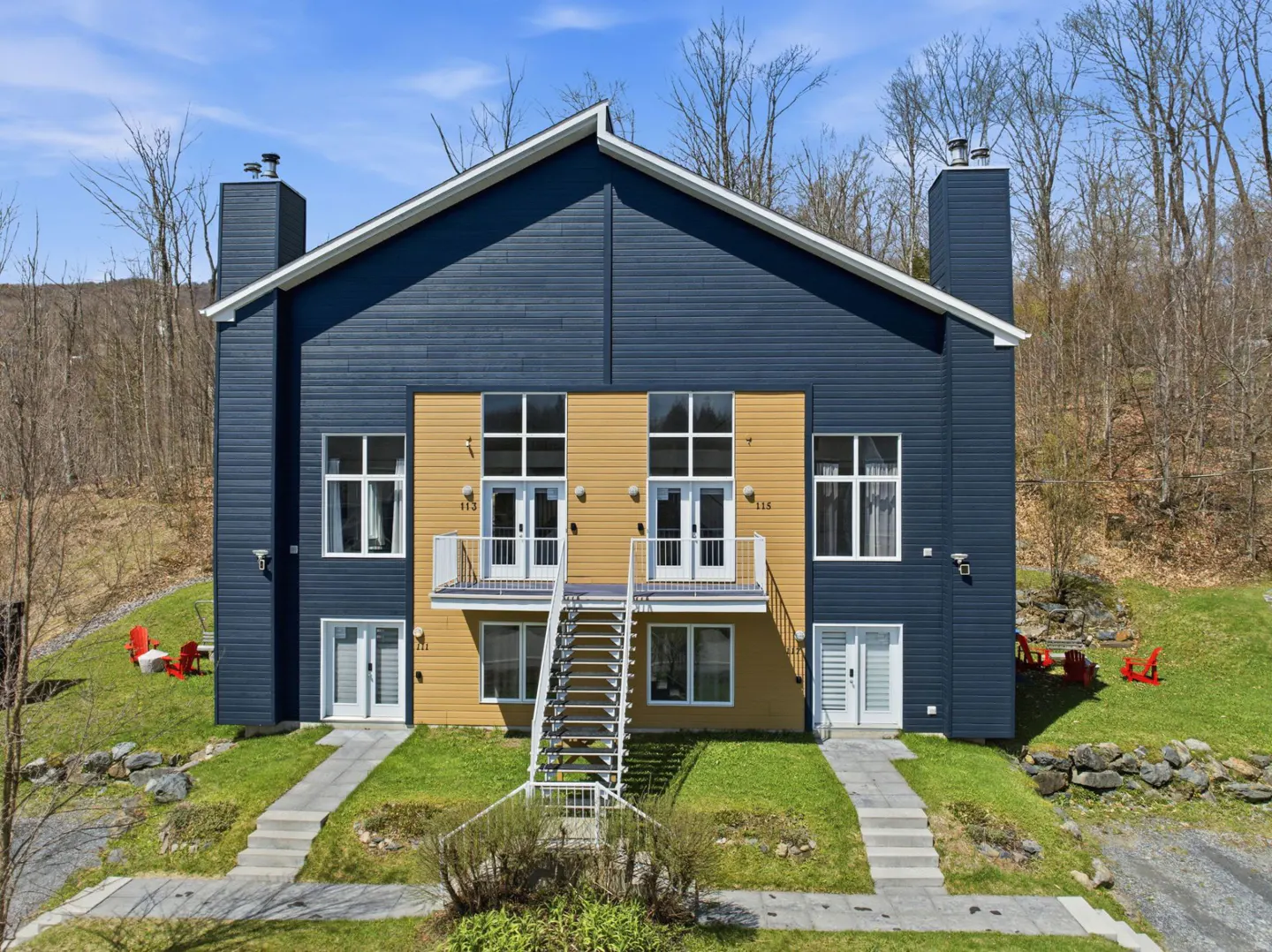 Two-story duplex with blue and yellow siding, white trim, and a shared staircase leading to upper balconies. Red chairs sit on the lawn.