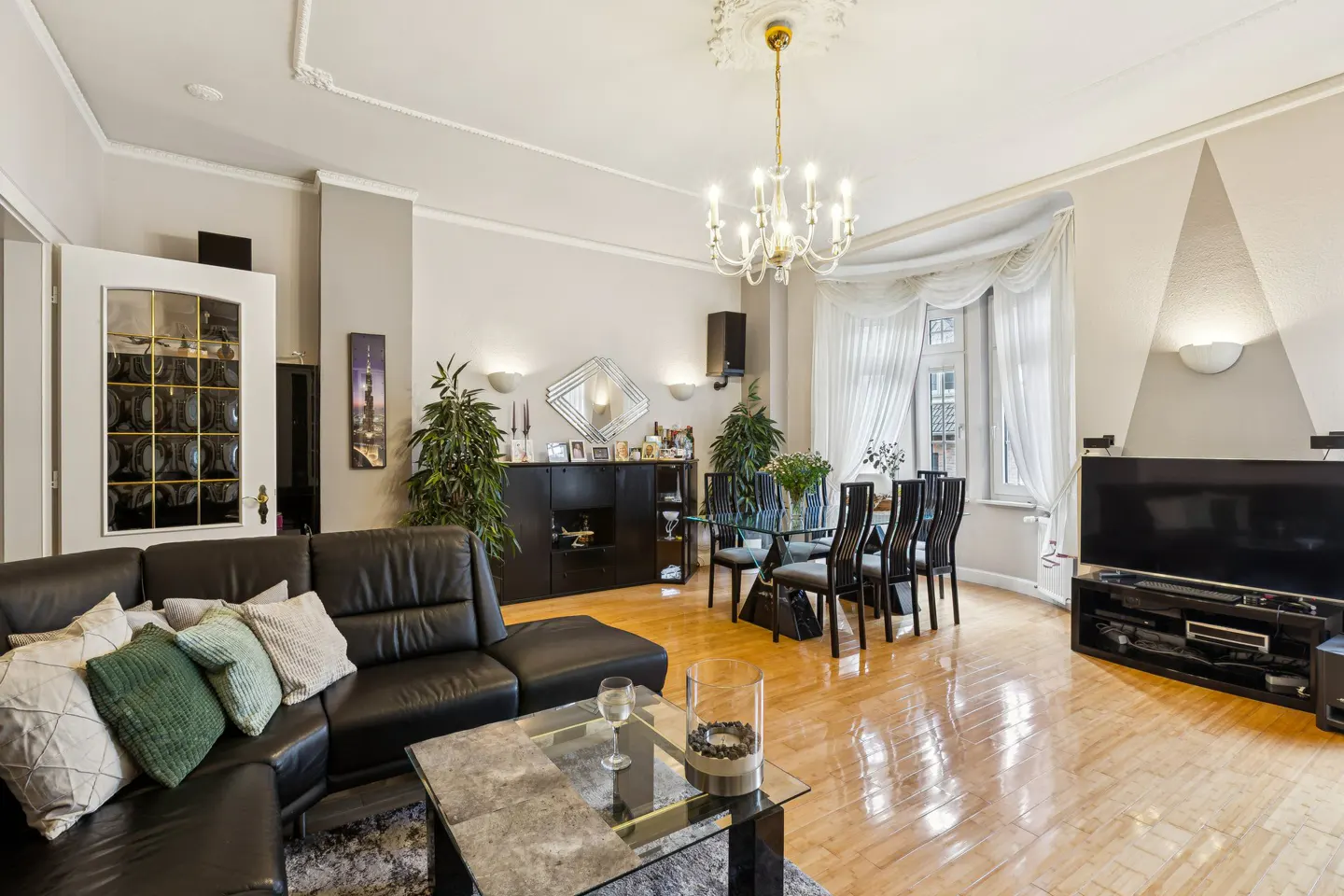 A living room with a black leather sofa, glass table, dining table, and a chandelier.