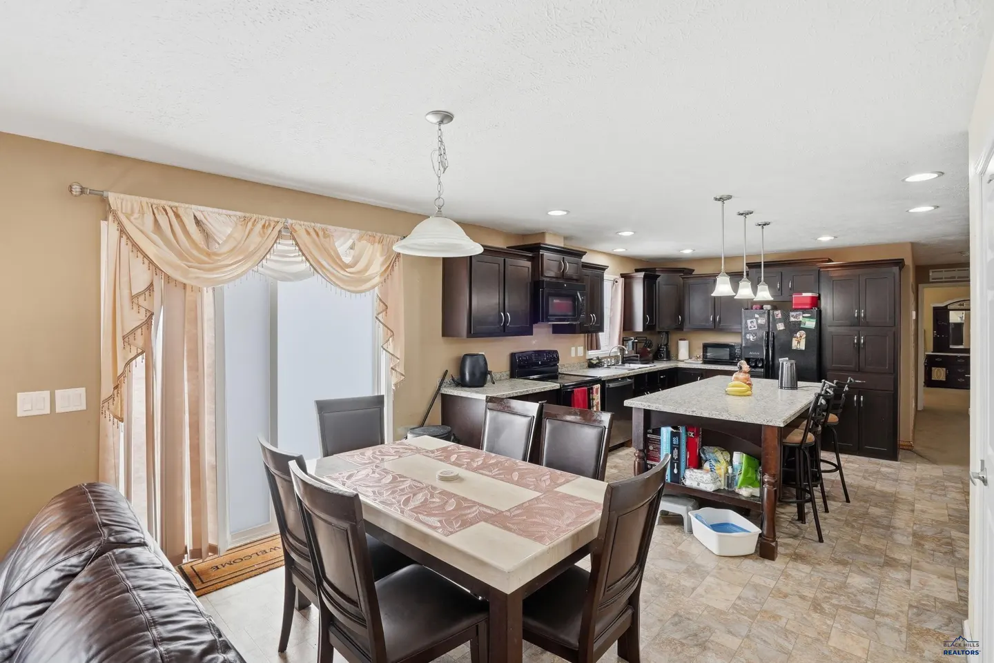 Open-concept kitchen and dining area with dark wood cabinets, island, and dining table with six chairs. Beige walls and tile flooring.