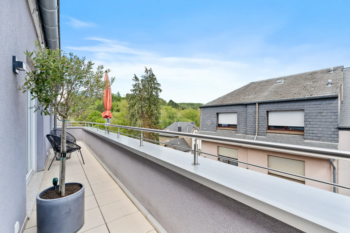 A long balcony with a potted tree, chairs, and a view of a house and trees.