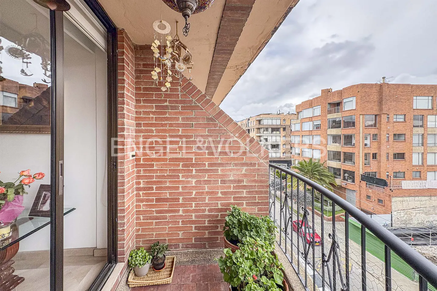 Balcony view with brick wall, plants, and black railing overlooking a city street with brick buildings under a cloudy sky.