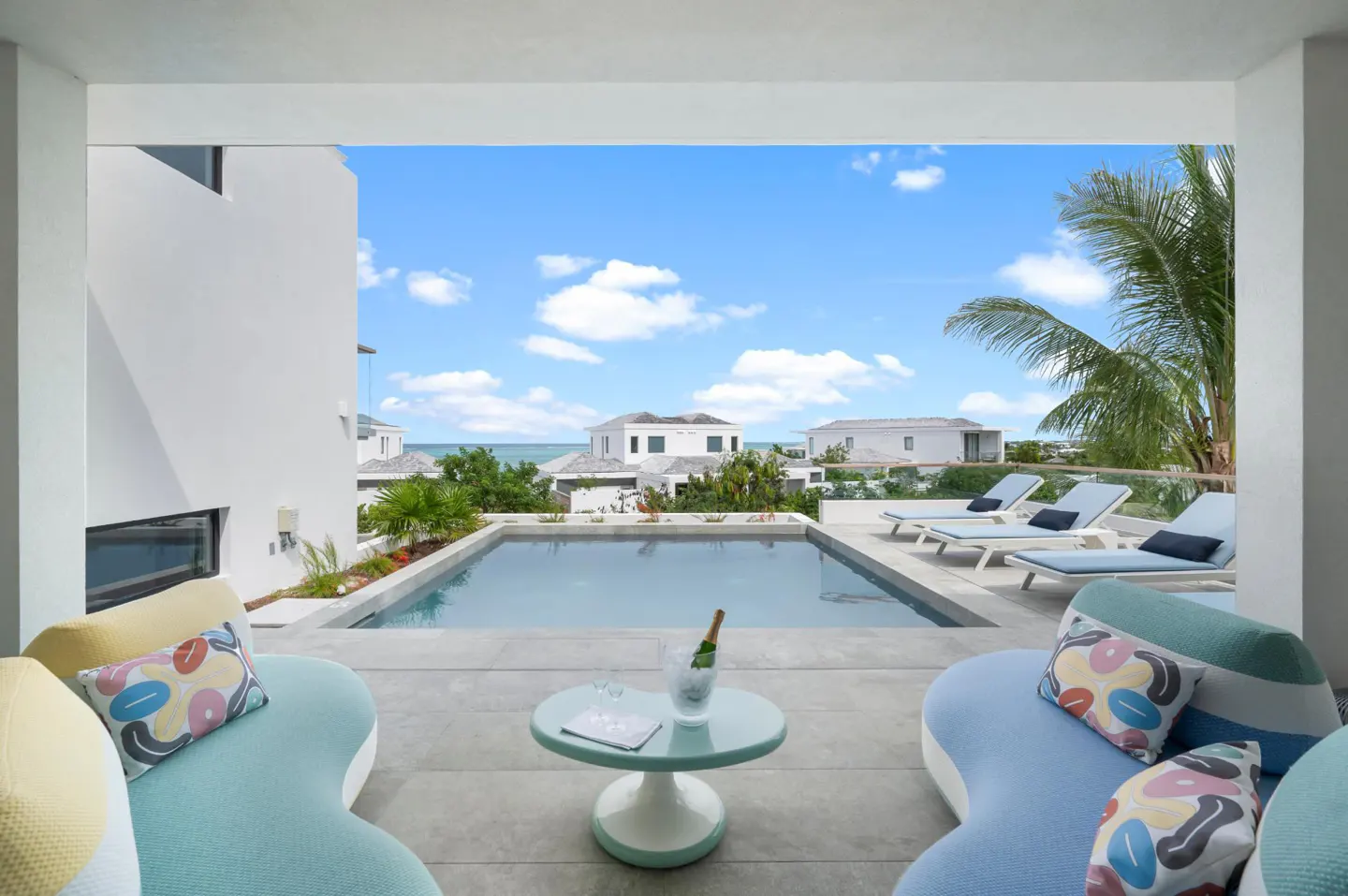 Luxury patio with blue couches, a pool, and ocean views. A bottle of champagne sits on a table. Blue sky with clouds.