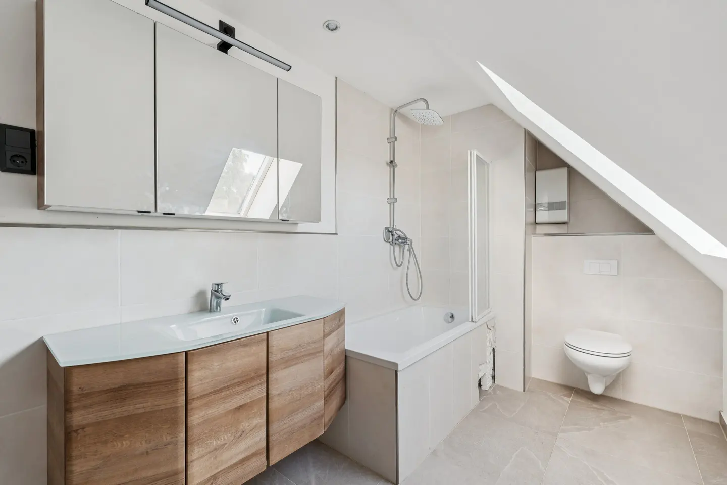Bright bathroom with a wood vanity, white sink, tub/shower, and toilet under a sloped ceiling.