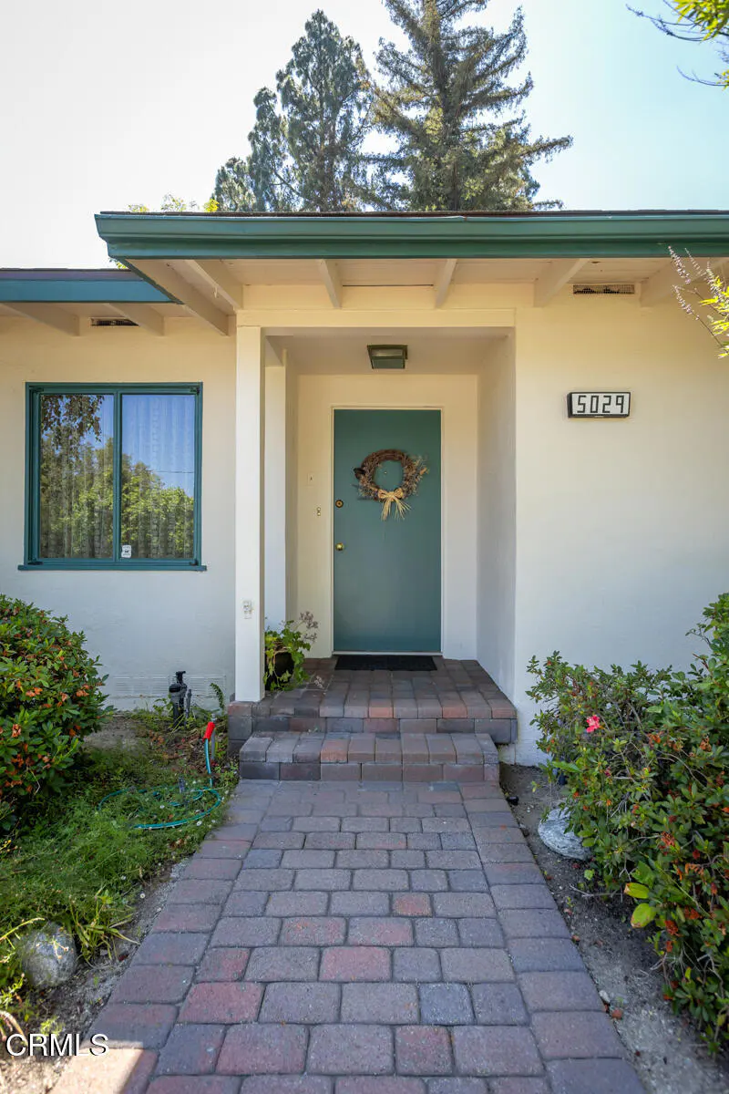 Front of a white house with a teal door and green trim. A brick path leads to the door, adorned with a wreath. Bushes line the walkway.
