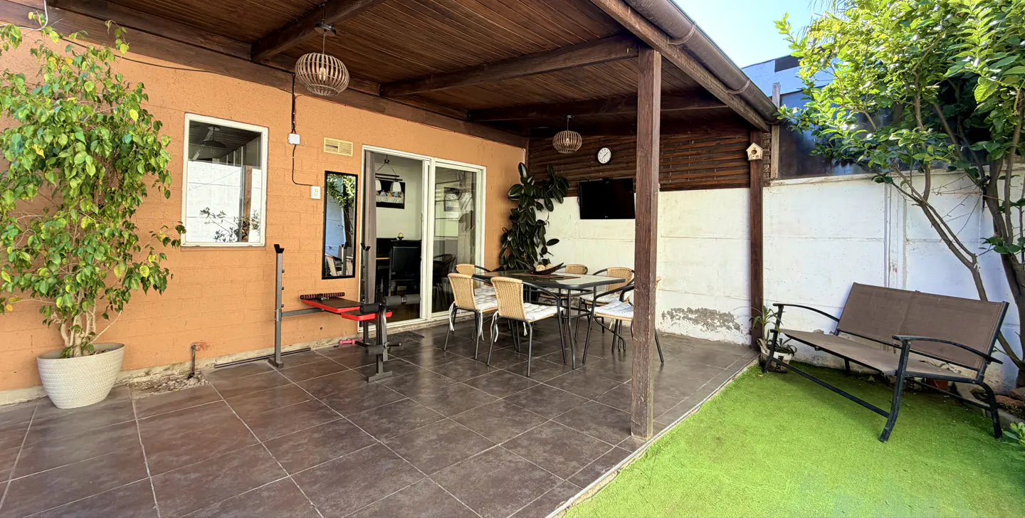 Outdoor patio with a wooden pergola, dining table, bench, and exercise equipment. The patio has brown tile flooring and a small patch of artificial grass.