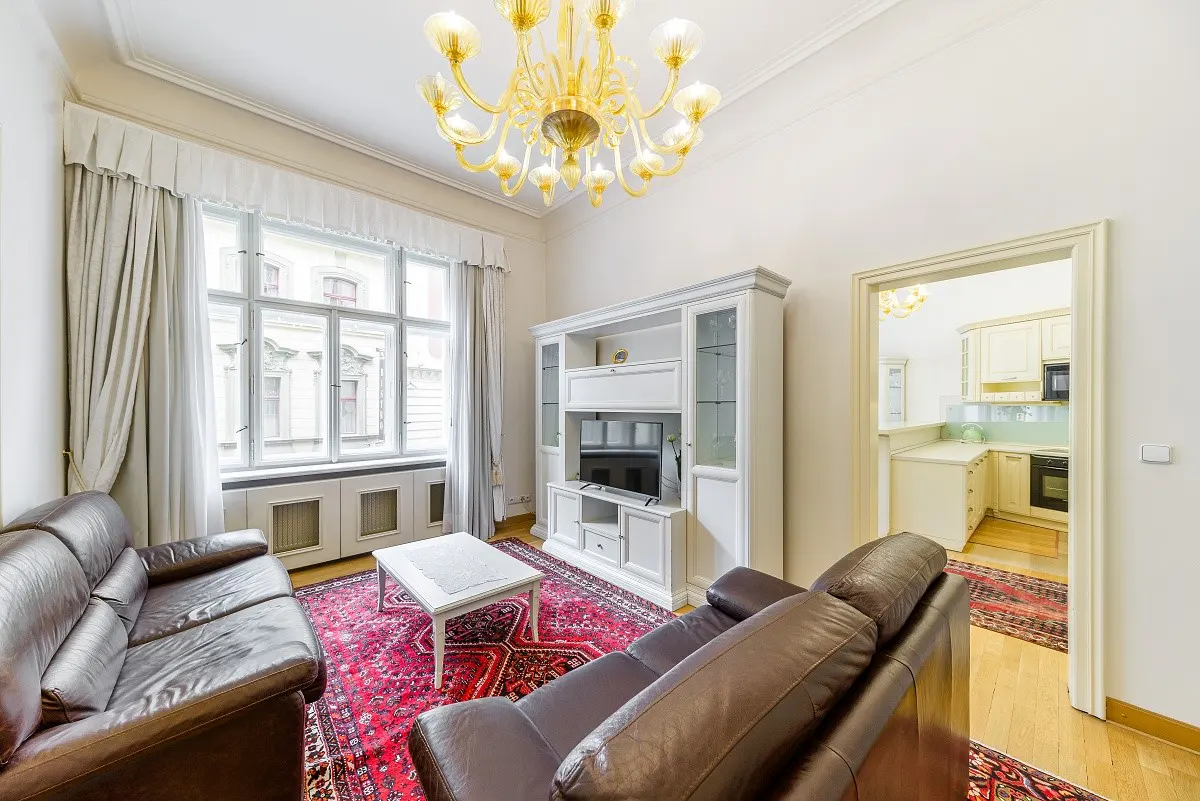 Living room with brown leather sofas, a red rug, and a white entertainment center with a TV. A doorway leads to a white kitchen. A gold chandelier hangs from the ceiling.