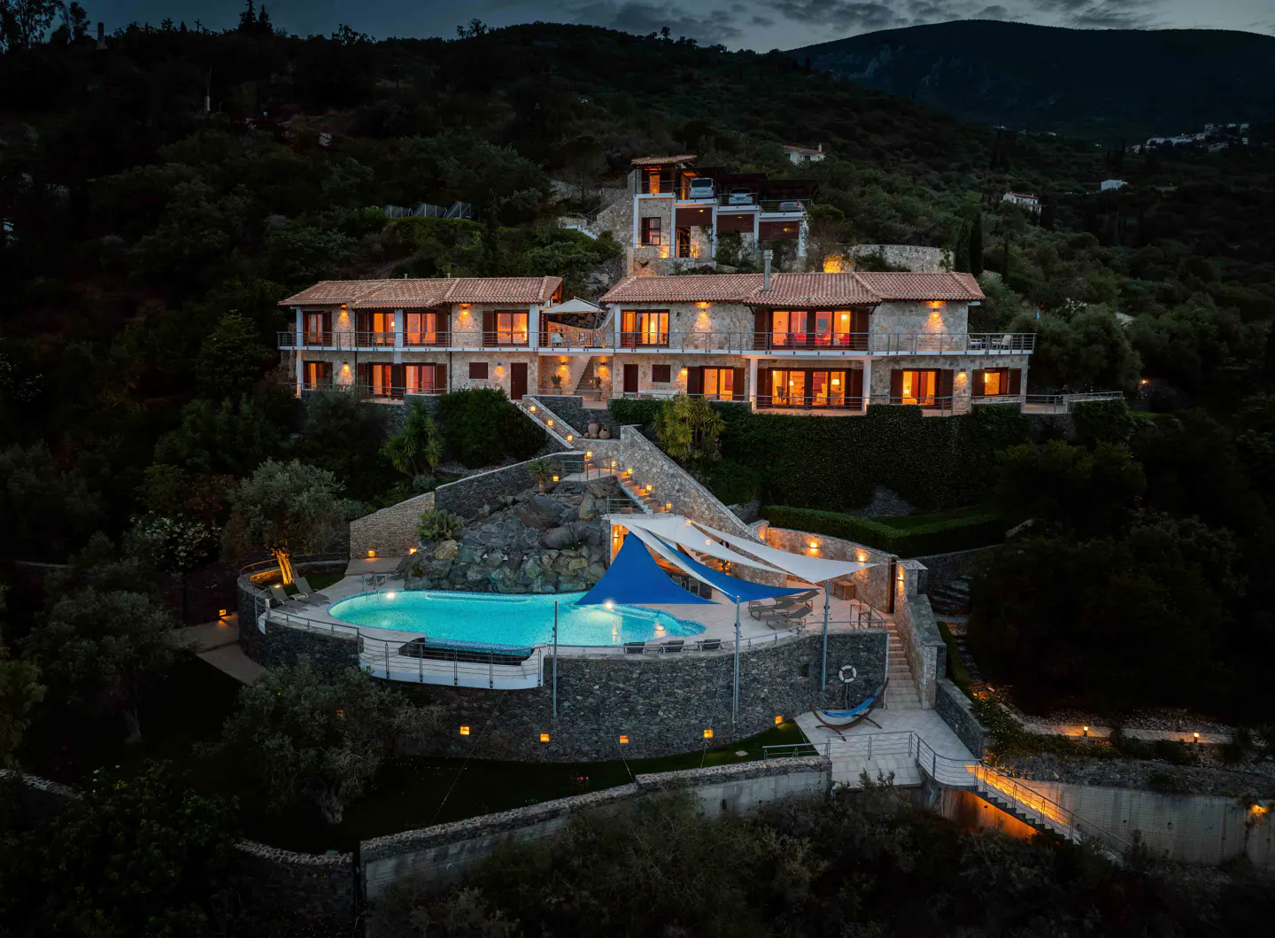 Night view of a stone villa with a red tile roof, a pool with blue canopies, and steps leading up a green hillside.