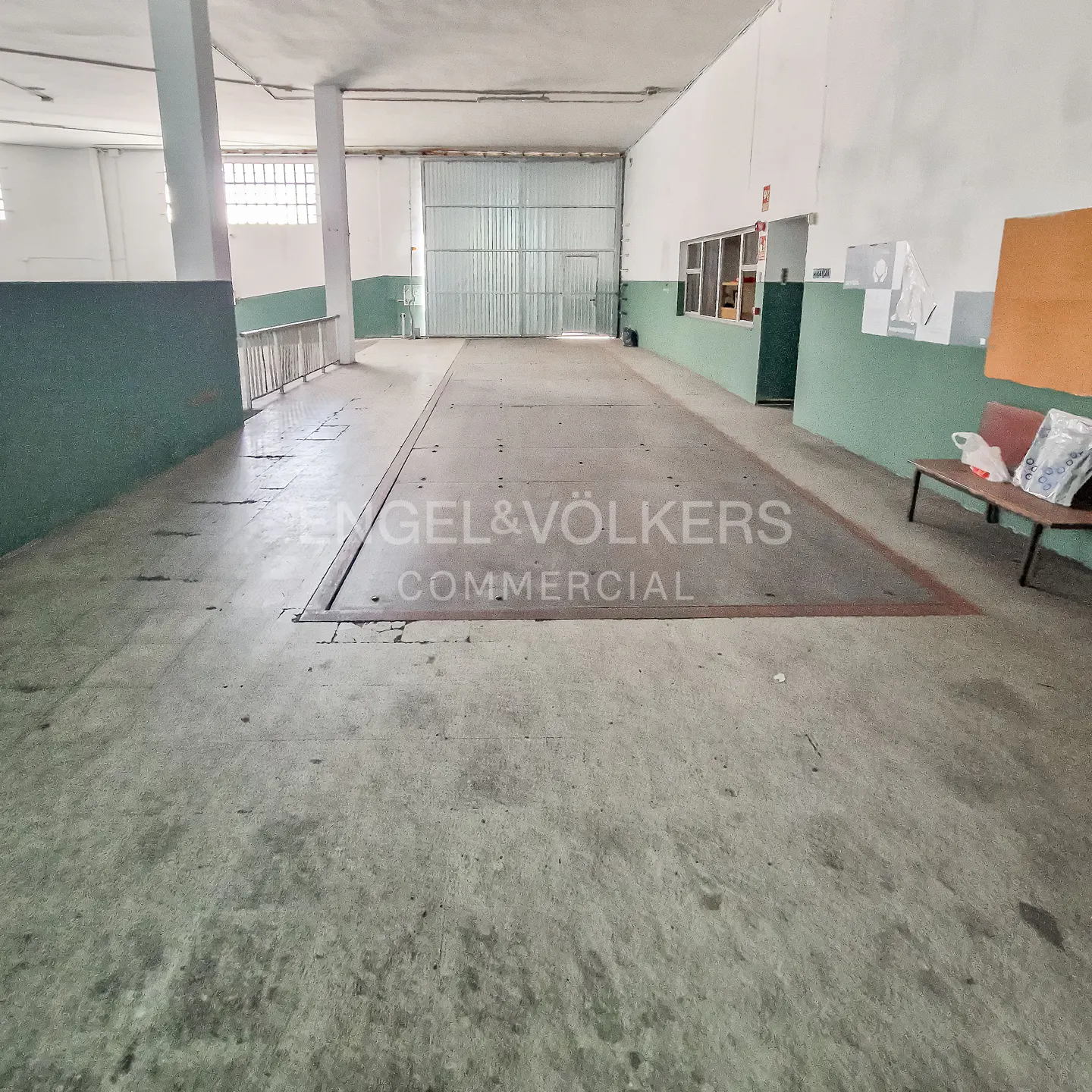 A wide shot of a large, empty warehouse with a concrete floor and a metal roll-up door. The walls are white with a green stripe. "Engel & Völkers Commercial" is printed on the floor.