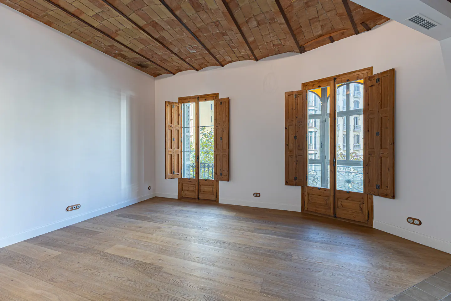 Empty room with wood floor, white walls, and brick ceiling. Two sets of wood doors with shutters open to city views.