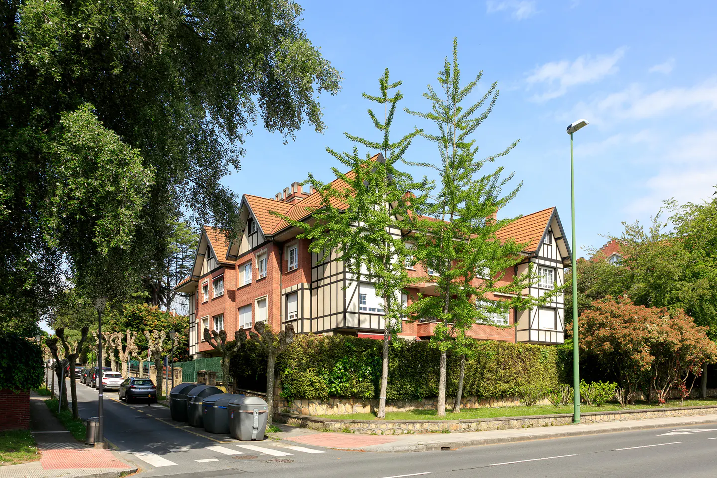 Brick apartment building with a red tile roof and Tudor-style accents, viewed from across a street with trees.