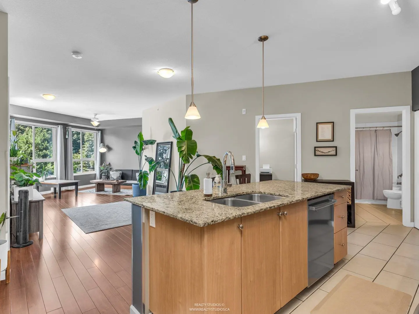 Open-concept living space with a kitchen island, stainless steel sink, granite countertop, and wood cabinets. Living room with large windows and plants.