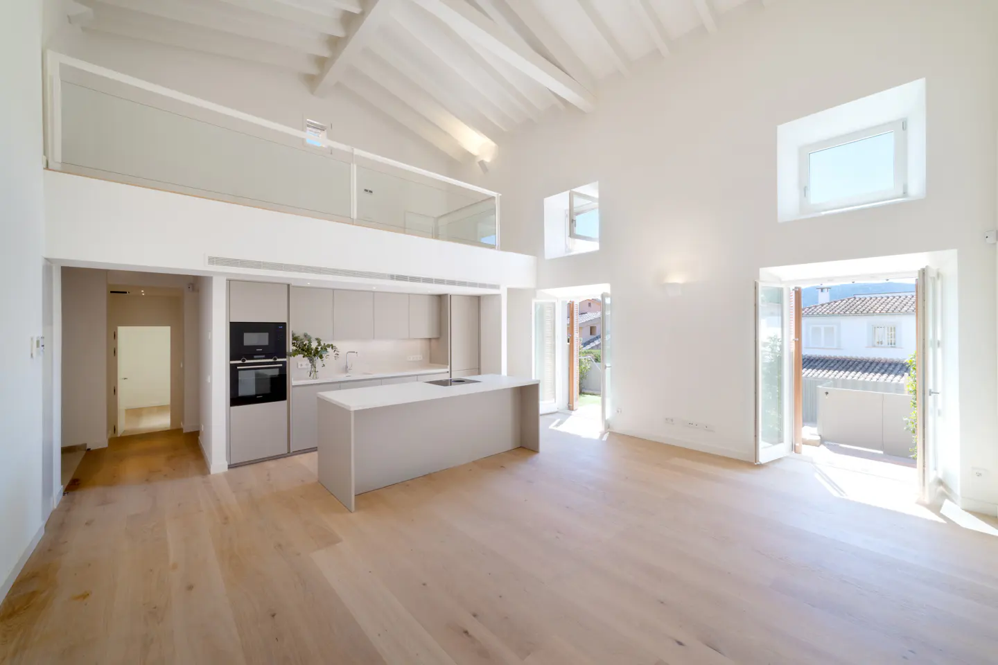 Bright, open-plan living space with white walls, wood floors, and a modern kitchen. French doors lead to an outdoor area. A loft overlooks the main floor.