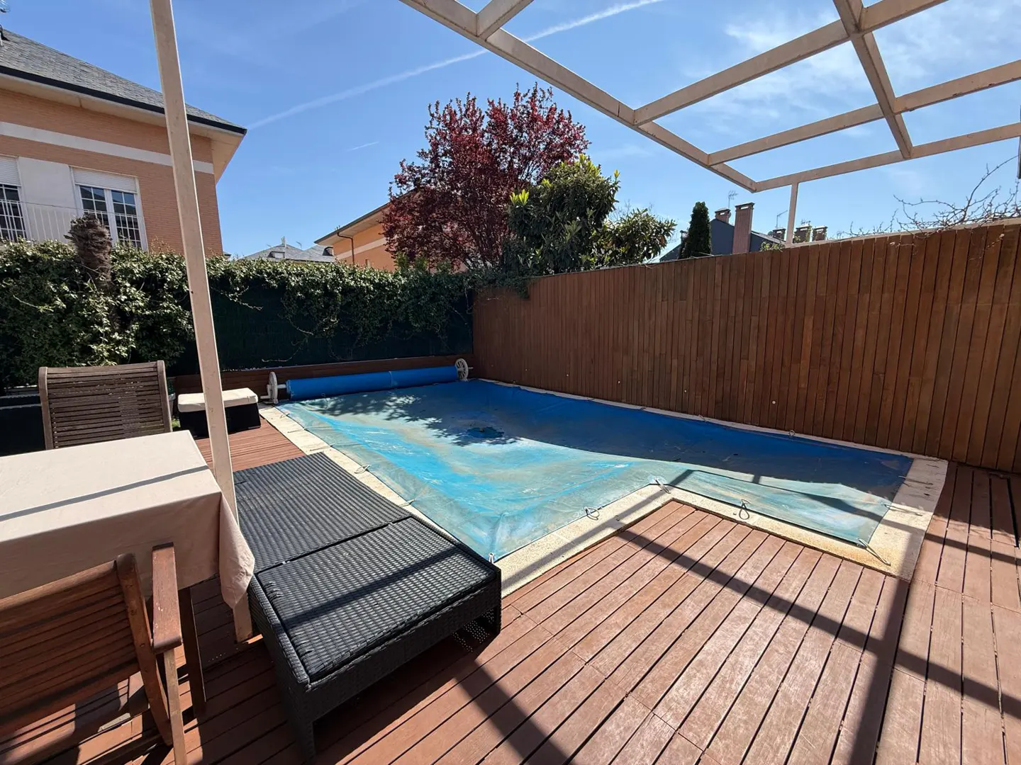Outdoor pool with a blue cover on a wooden deck, surrounded by a wooden fence and greenery. Table and chairs are on the left.