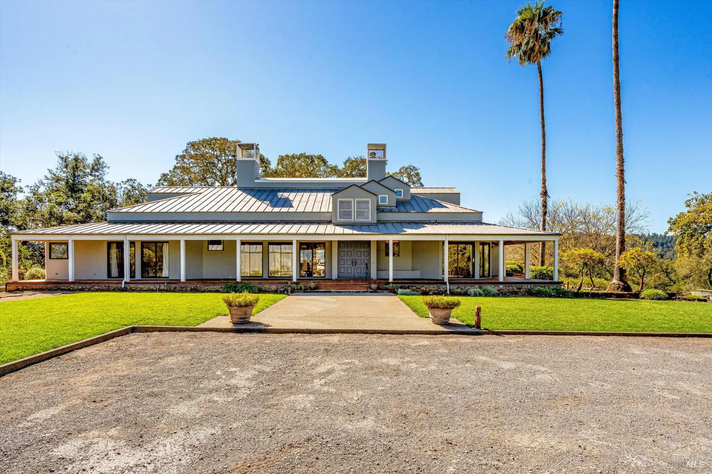 Wide shot of a one-story gray house with a metal roof, white pillars, and a gravel driveway on a sunny day.