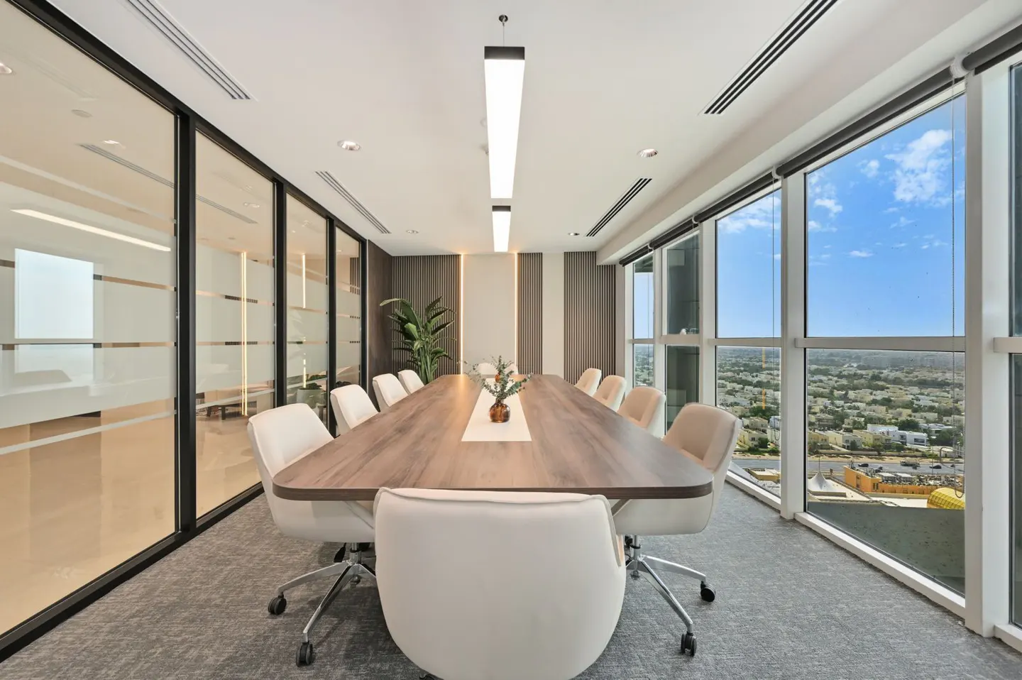 Bright, modern conference room with a long wood table, white chairs, and floor-to-ceiling windows overlooking a city.