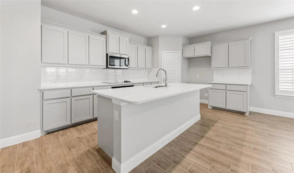 Bright kitchen with gray cabinets, white countertops, and wood-look tile flooring. A large island with a sink is in the center.