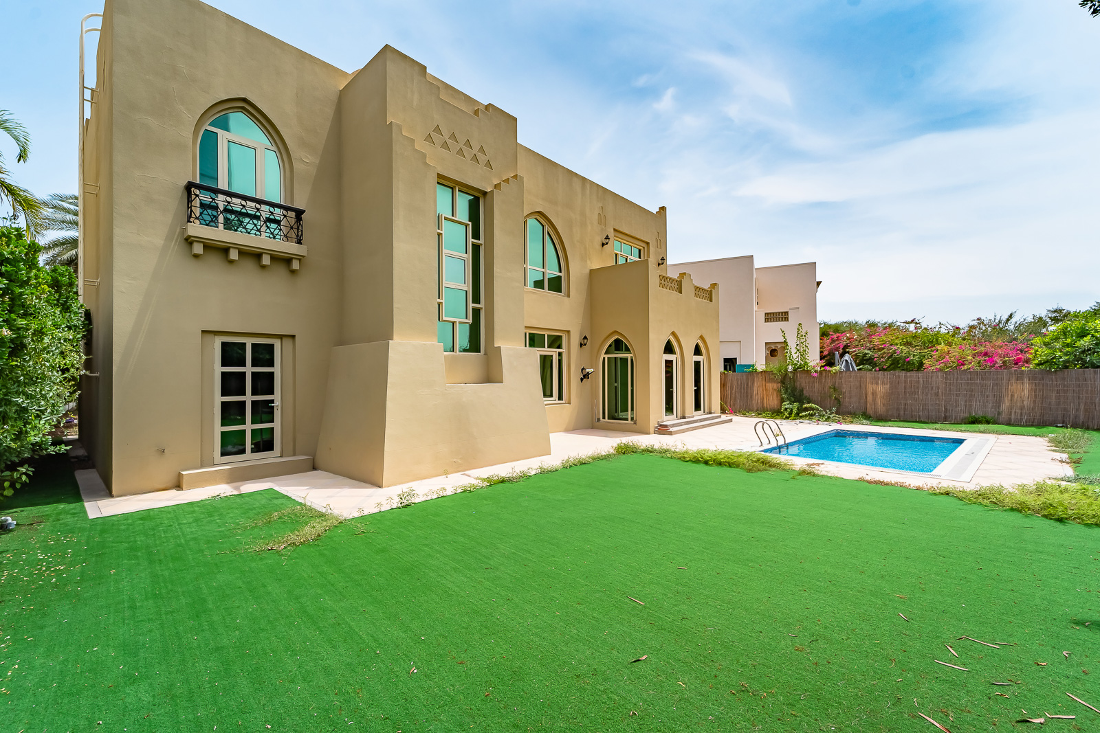 Tan villa with arched windows and a balcony, featuring a green lawn and a blue swimming pool under a bright blue sky.