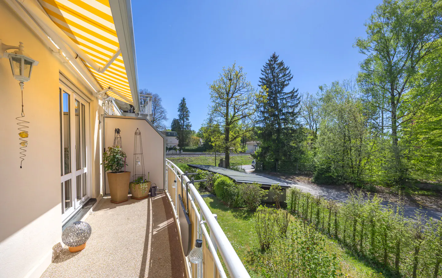 Balcony view with yellow striped awning, white railing, and potted plants. Trees and blue sky in the background.