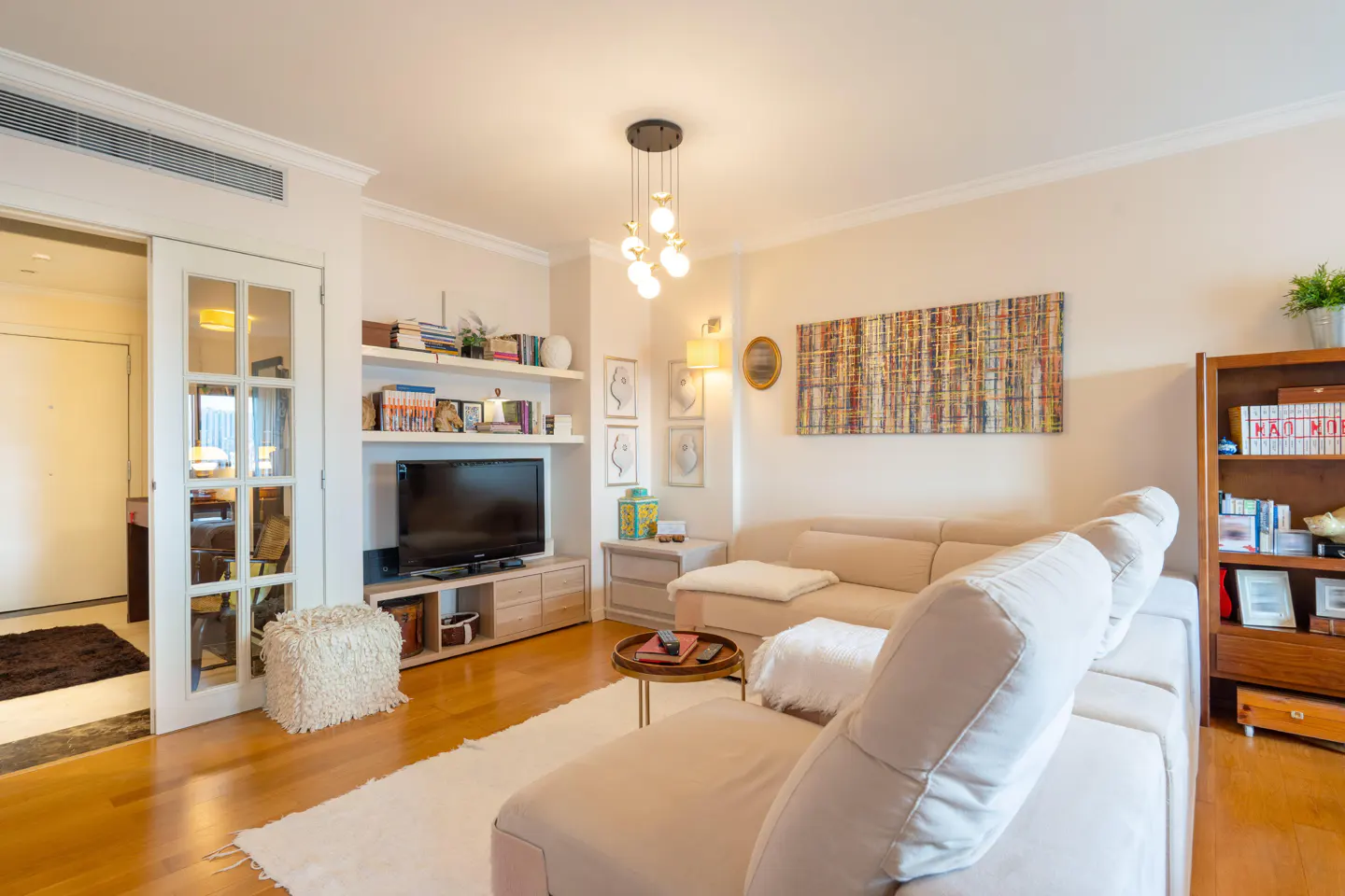 A bright living room with a beige sectional sofa, wood floors, TV, and bookshelves. A white rug and modern light fixture add to the room's appeal.