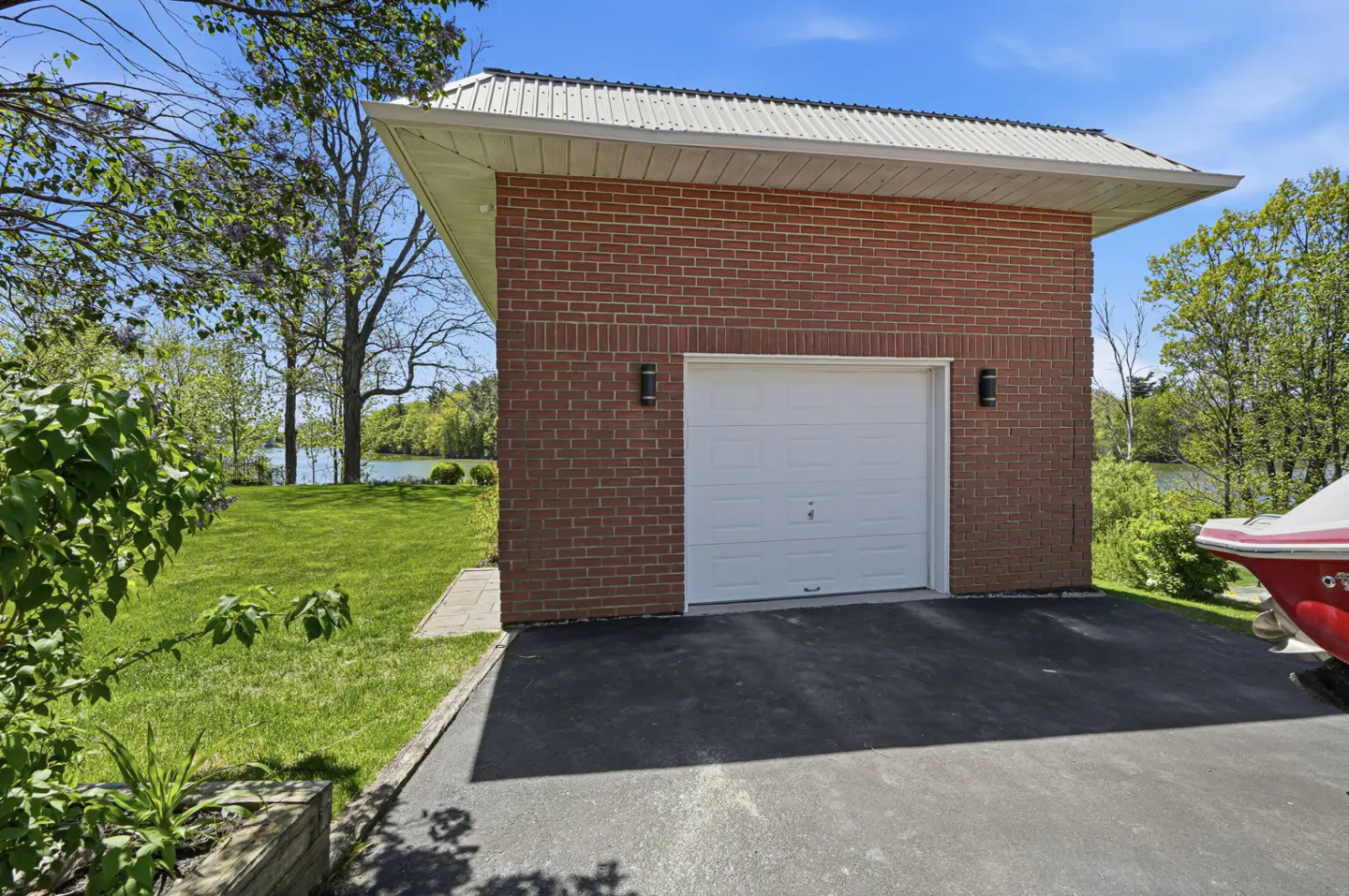 A brick garage with a white door and metal roof sits on a black asphalt driveway near a lake.