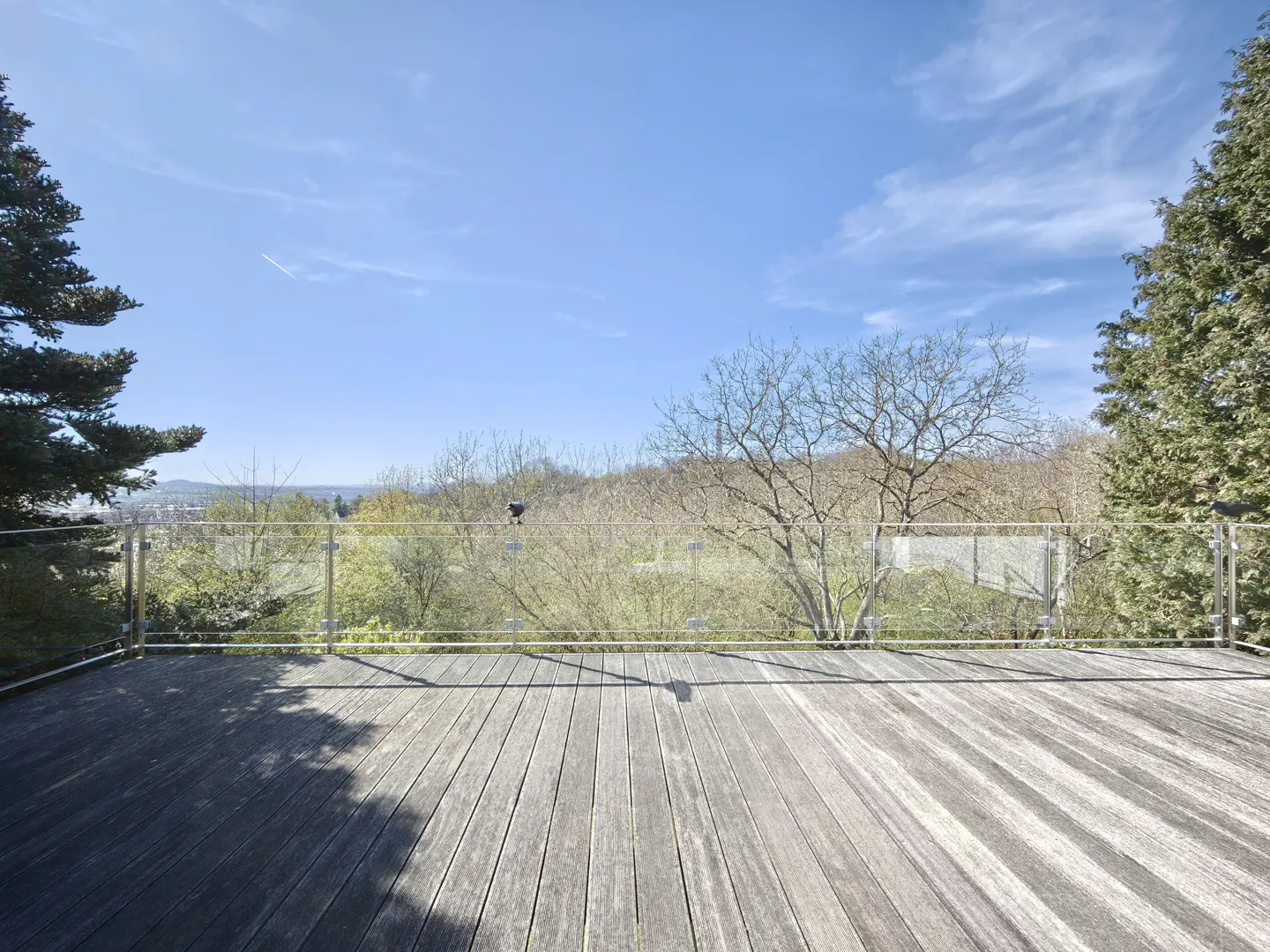 Wide shot of a wooden deck with a glass railing overlooking trees and a blue sky.