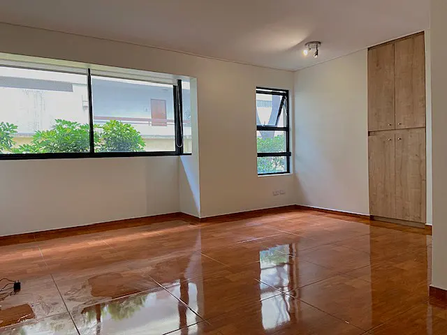 Bright, empty room with shiny wood floors, white walls, and two windows. A tall, light-brown wooden cabinet stands against the right wall.