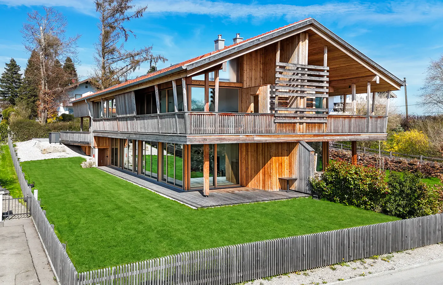 Two-story wood house with a balcony, large windows, and a green lawn enclosed by a wooden fence under a blue sky.