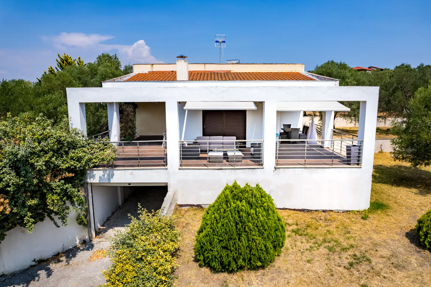 White two-story house with a red tile roof, a balcony with furniture, and a garage entrance, surrounded by green trees under a blue sky.