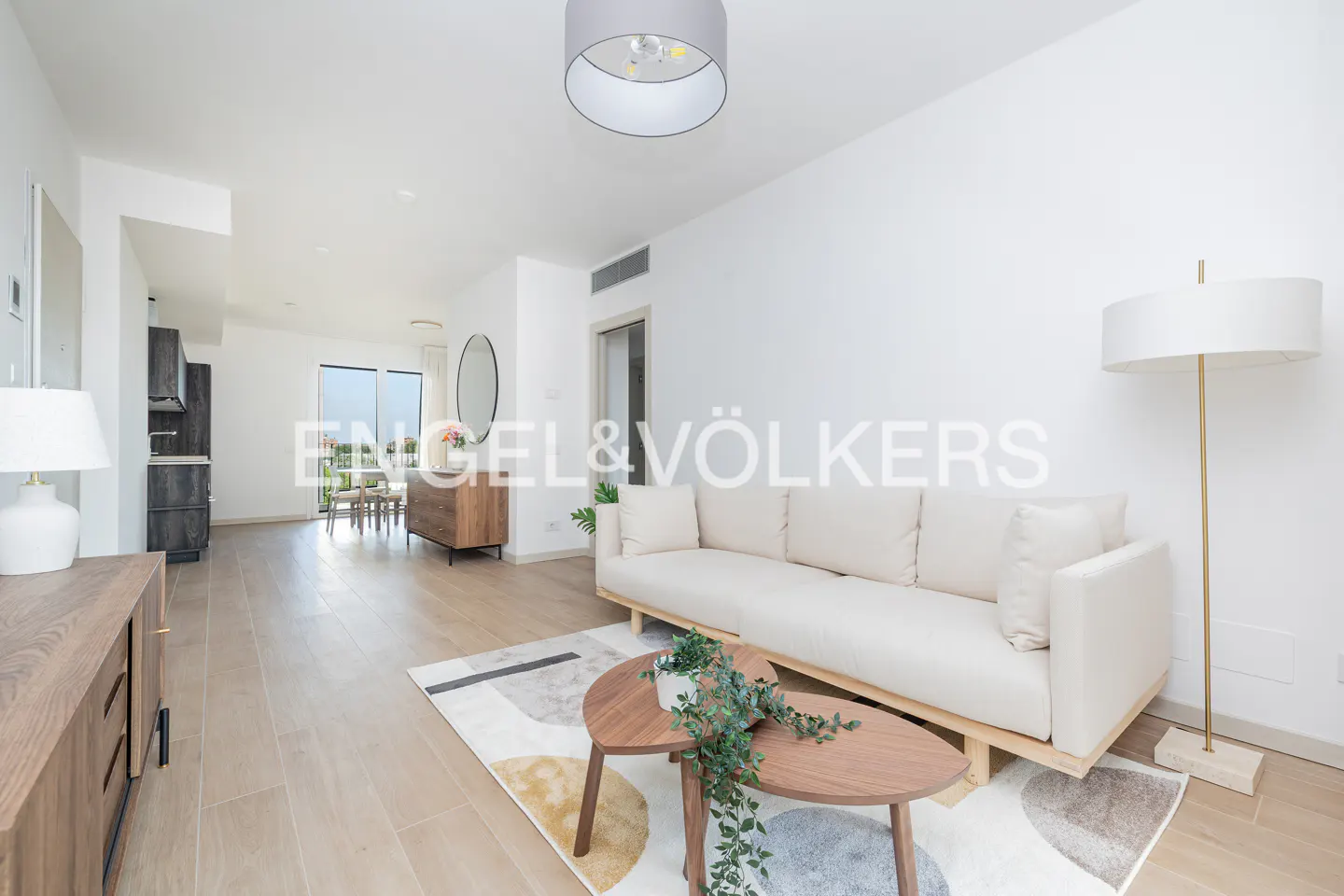 Bright living room with white walls, light wood floors, and a cream-colored sofa. A two-tiered coffee table sits on a patterned rug. Engel & Völkers logo visible.