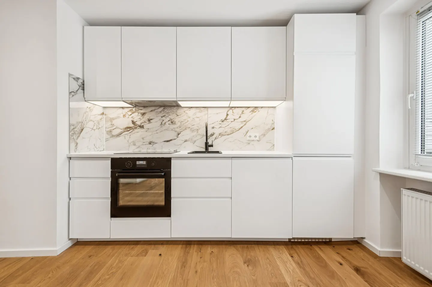 A modern kitchen with white cabinets, marble backsplash, black oven, and wood floor.