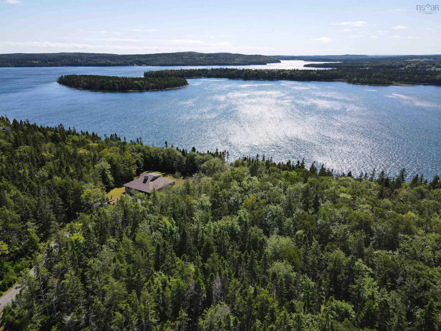 Aerial view of a brown house nestled in a green forest, overlooking a blue bay with islands under a clear sky.