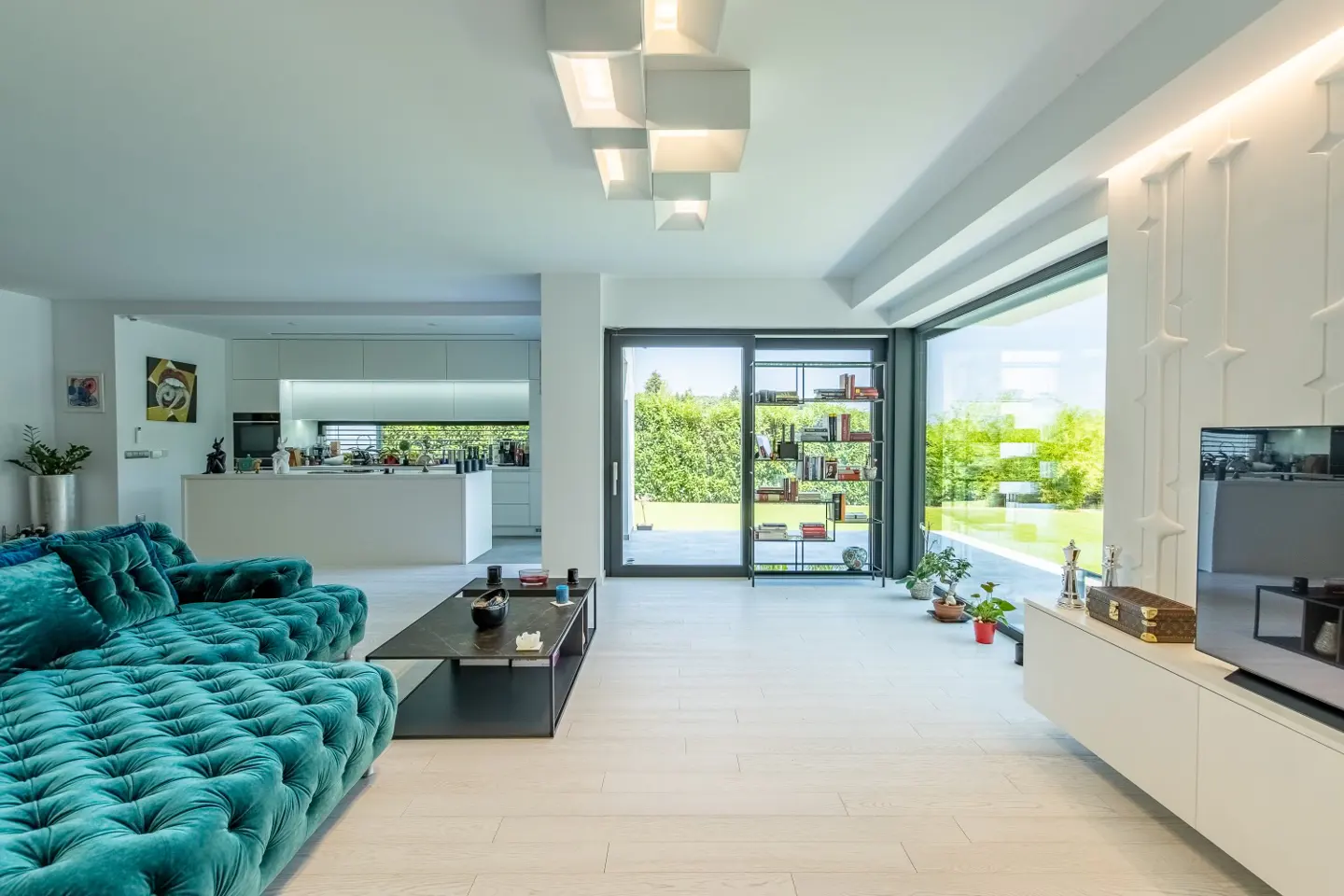 Bright, modern living room with a teal tufted sofa, black coffee table, and open kitchen. Large windows overlook a green lawn.
