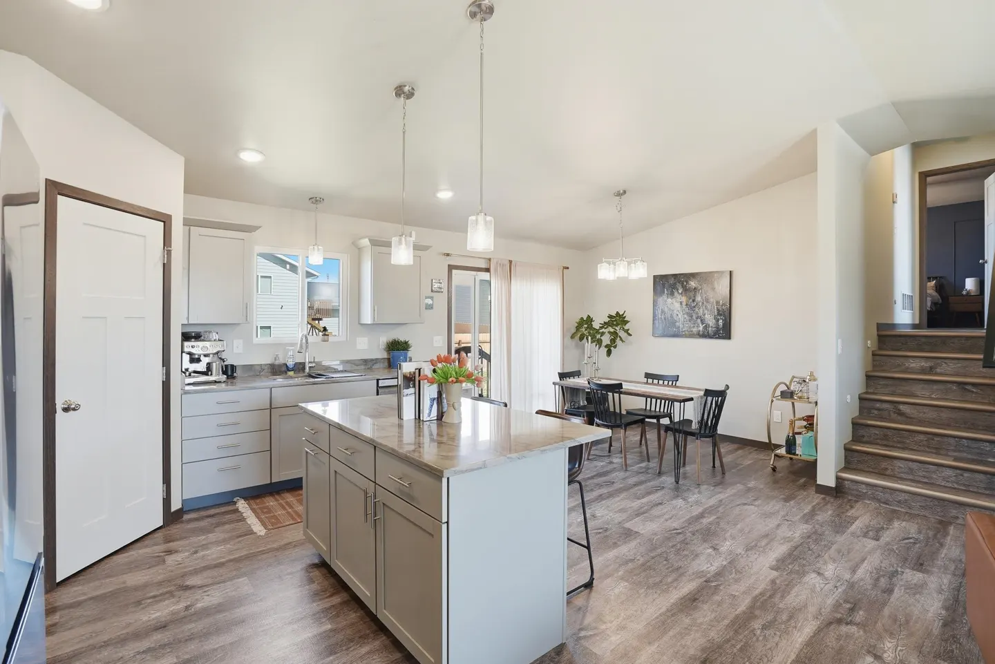 Open-concept kitchen and dining area with gray cabinets, island, wood floors, and staircase. Natural light fills the space.
