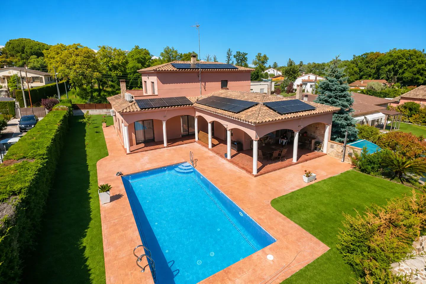 Aerial view of a salmon-colored house with solar panels, a pool, and a covered patio with white columns. Green lawn and trees surround the property.