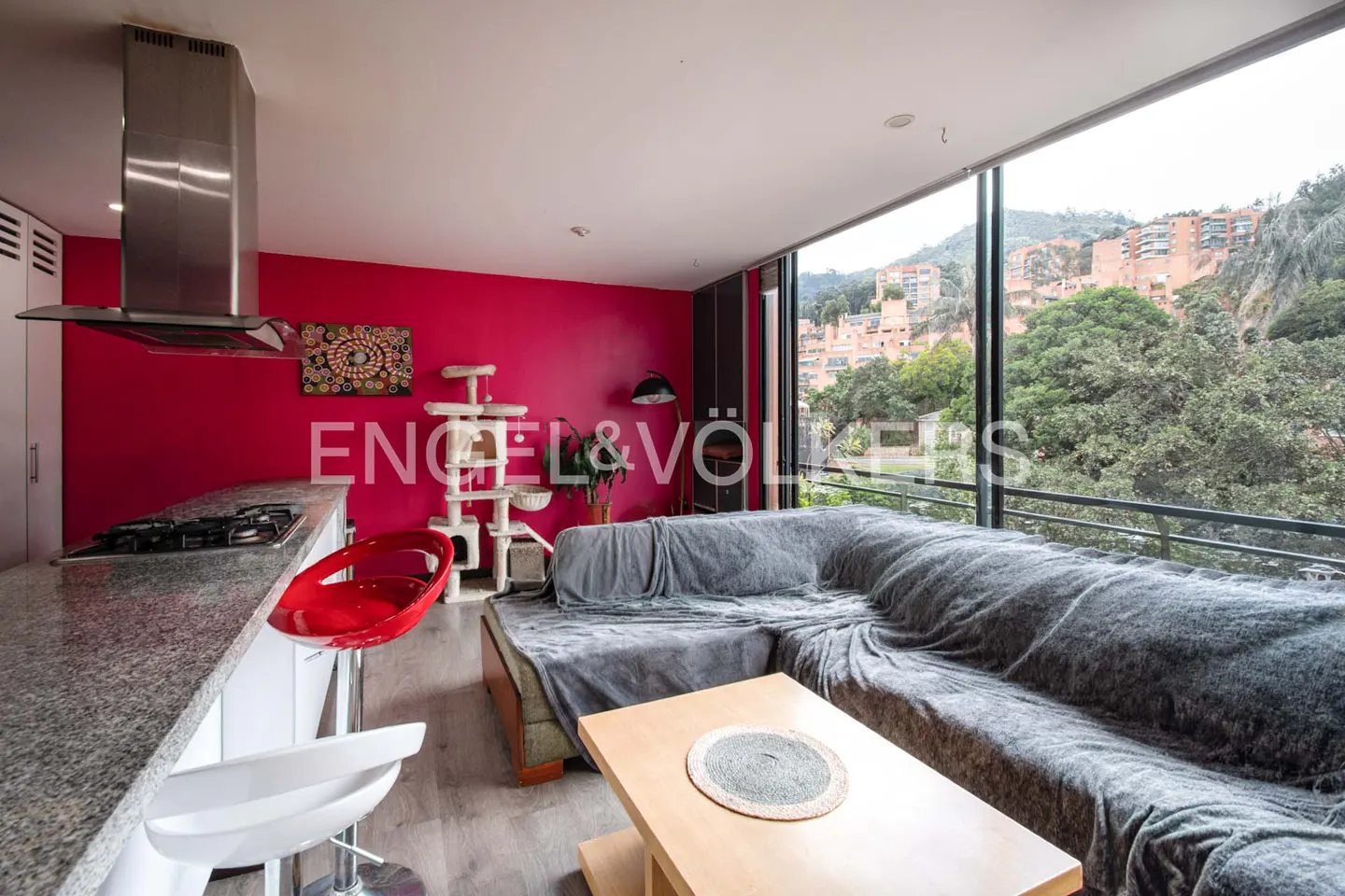Bright living room with pink wall, gray sectional sofa, and city view through large windows. Kitchen island with stools visible.