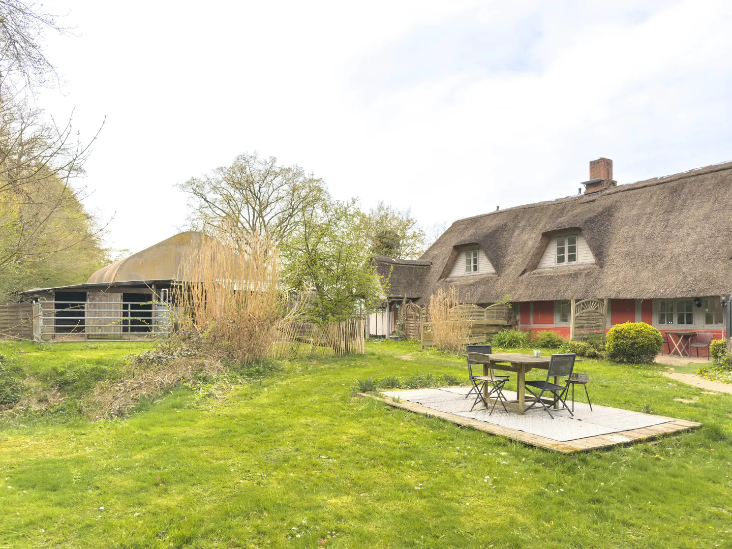 Exterior view of a red house with a thatched roof, a green lawn, and a table with chairs.