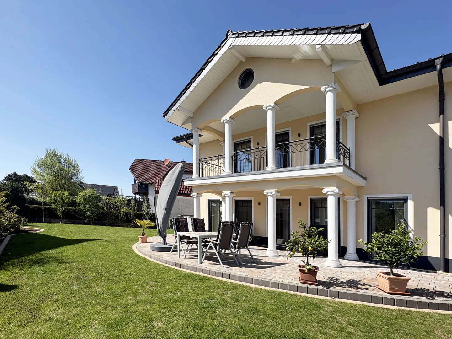 Two-story beige house with white columns and black trim. Patio with table, chairs, and potted plants. Green lawn and blue sky.