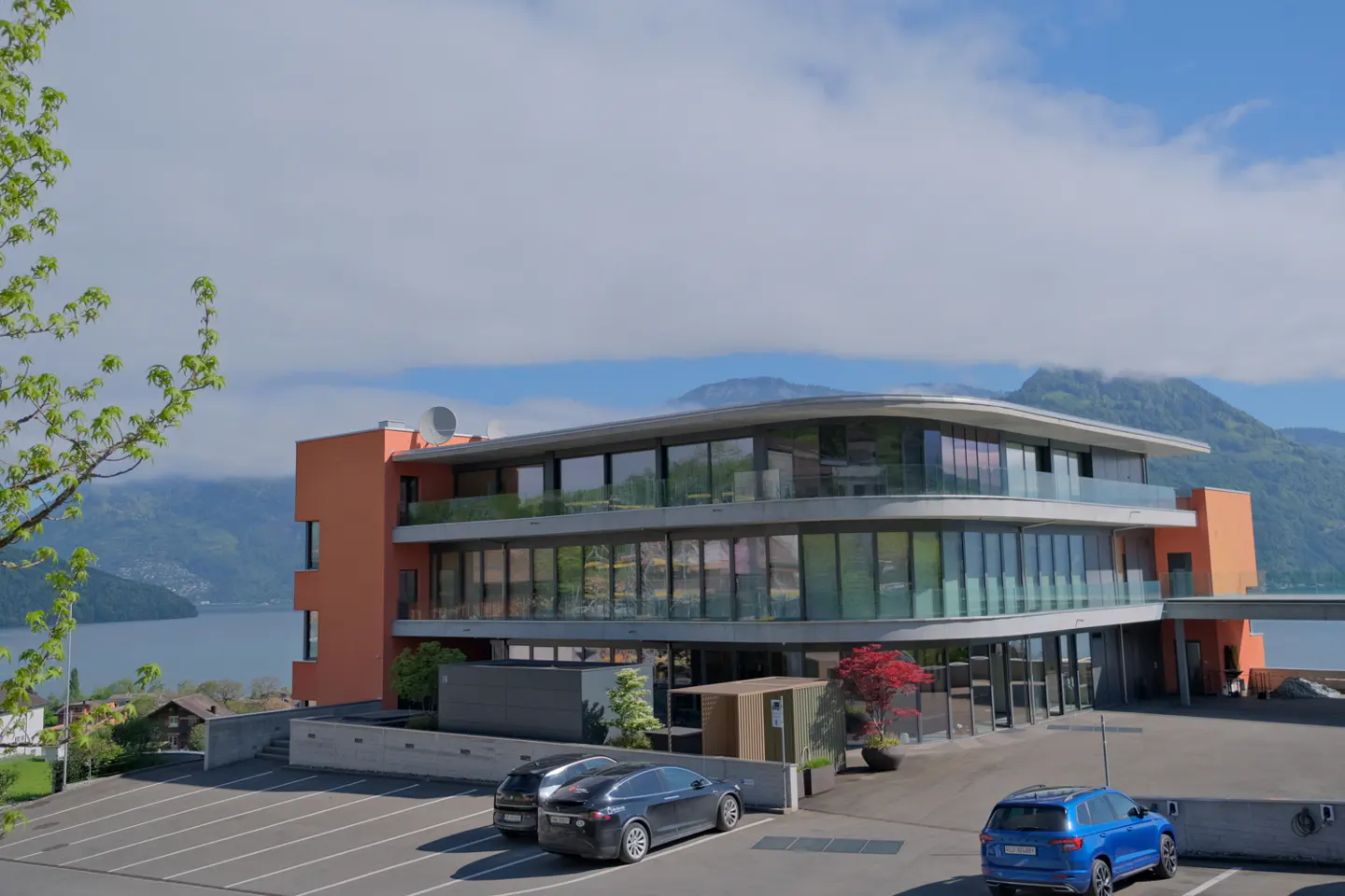 Modern orange building with glass windows, a parking lot with cars, and a lake and mountains in the background.