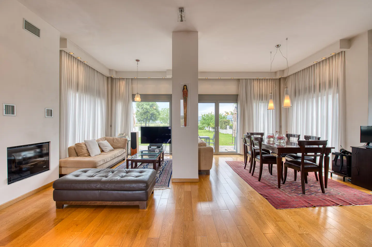 Open-concept living and dining room with hardwood floors, beige sofa, and dark wood dining table set. Large windows with sheer curtains.