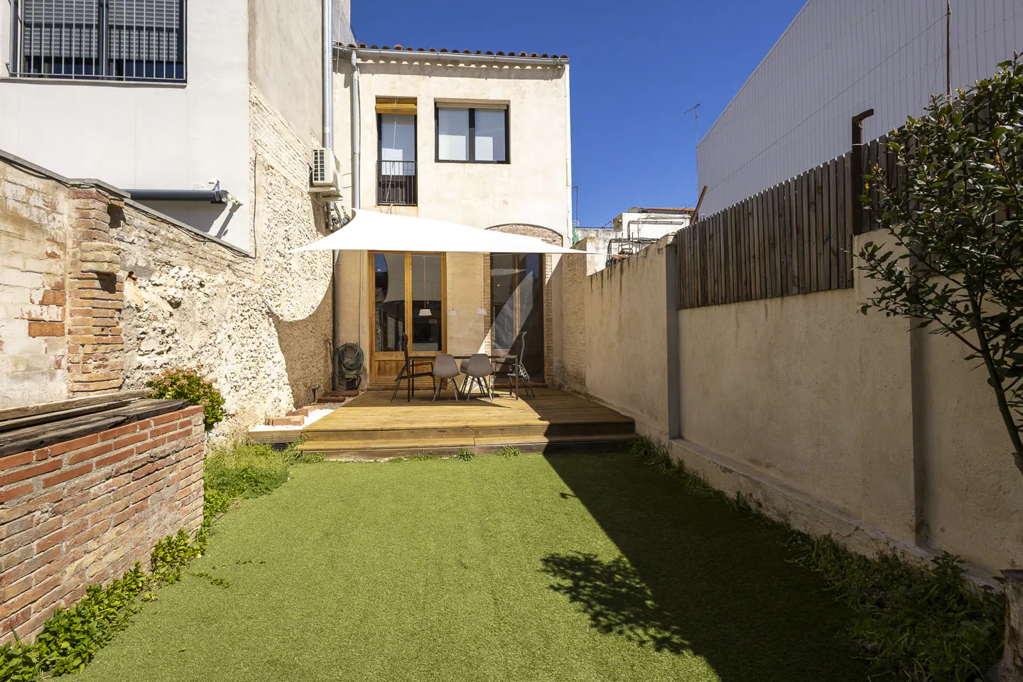 A backyard with a wooden deck, chairs, and a white sunshade. Green grass covers the ground, bordered by brick and stucco walls.