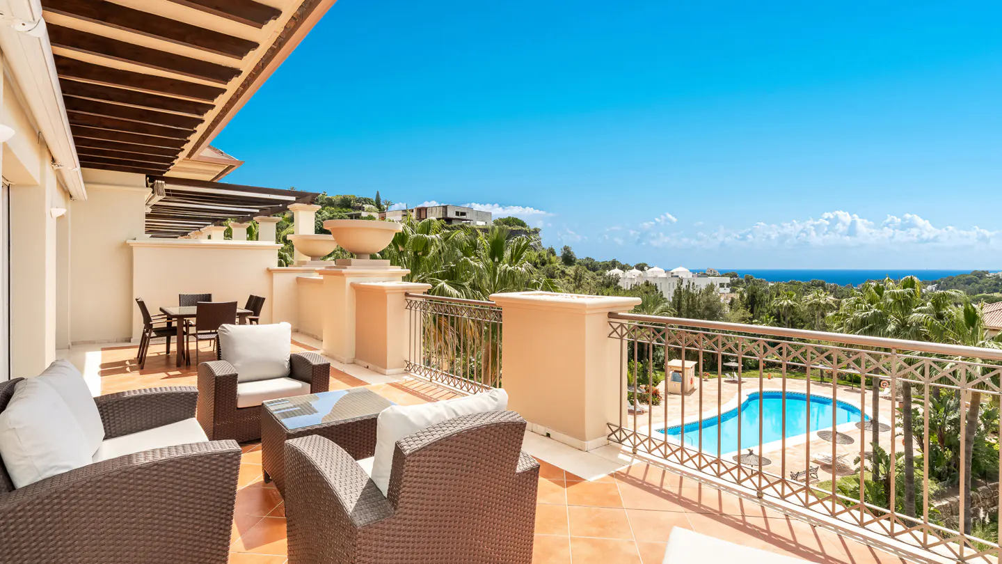 A sunny patio with wicker furniture overlooks a pool, palm trees, and the ocean under a clear blue sky.