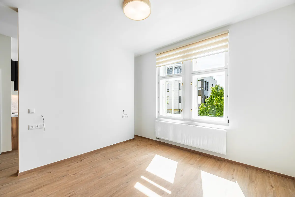 Bright, empty room with wood floors, white walls, and a large window with a view of trees and buildings. A light fixture is on the ceiling.