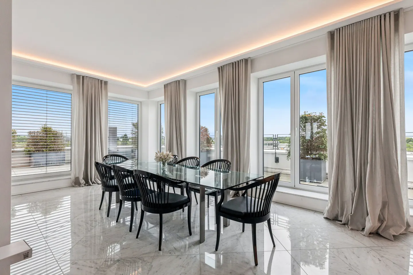 Bright dining room with marble floors, a glass table, and black chairs. Large windows with beige curtains offer a view of the outside.