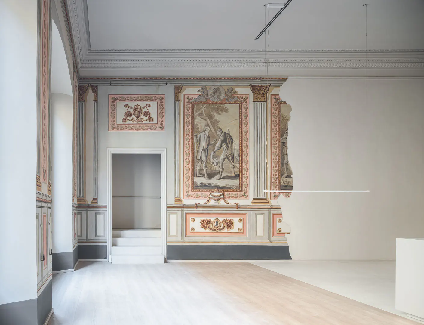 A room with ornate wall paintings, including figures, and a doorway leading to stairs. One wall is partially unfinished.