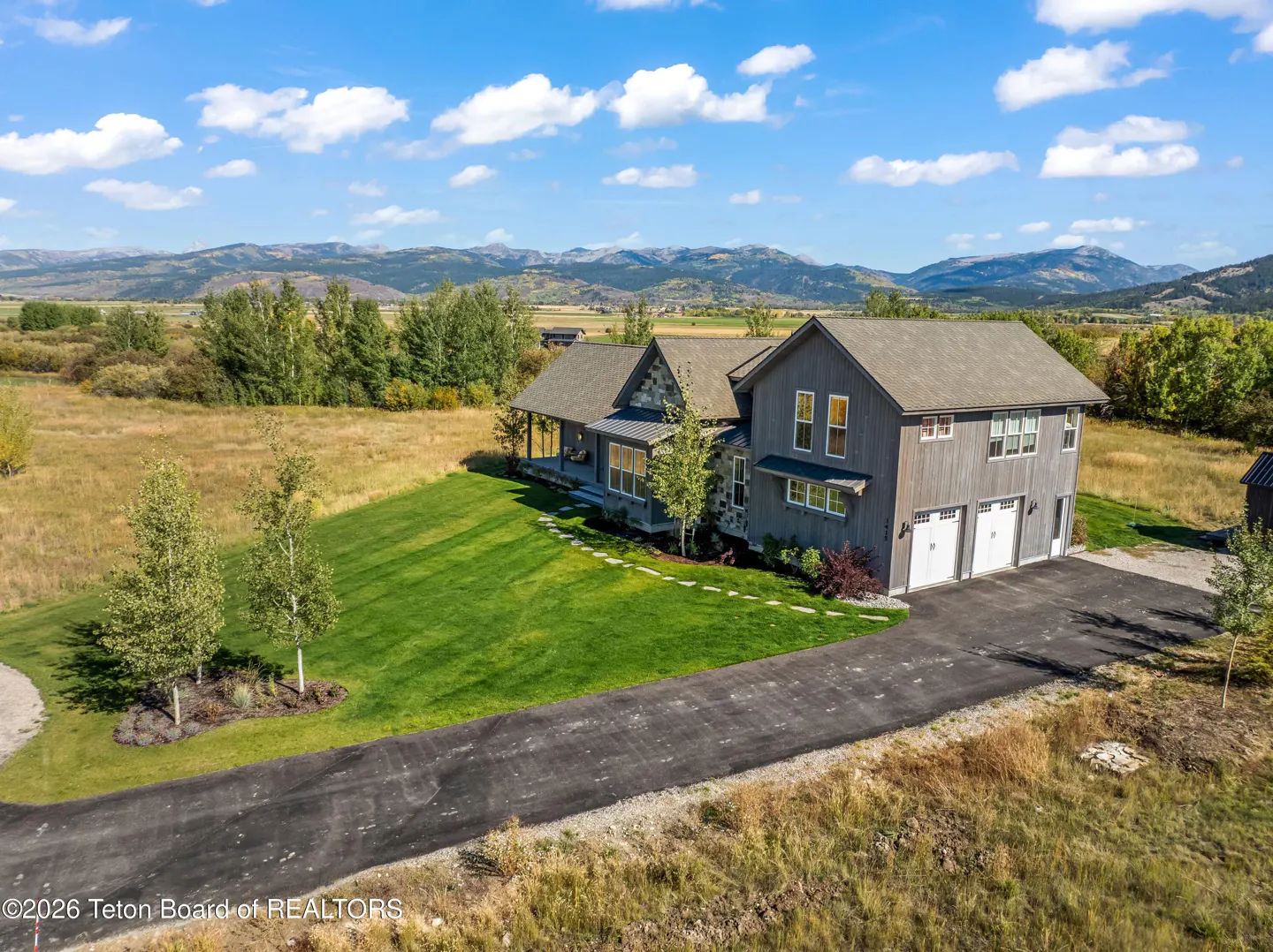 Aerial view of a gray two-story house with a green lawn, a black driveway, and mountains in the background.