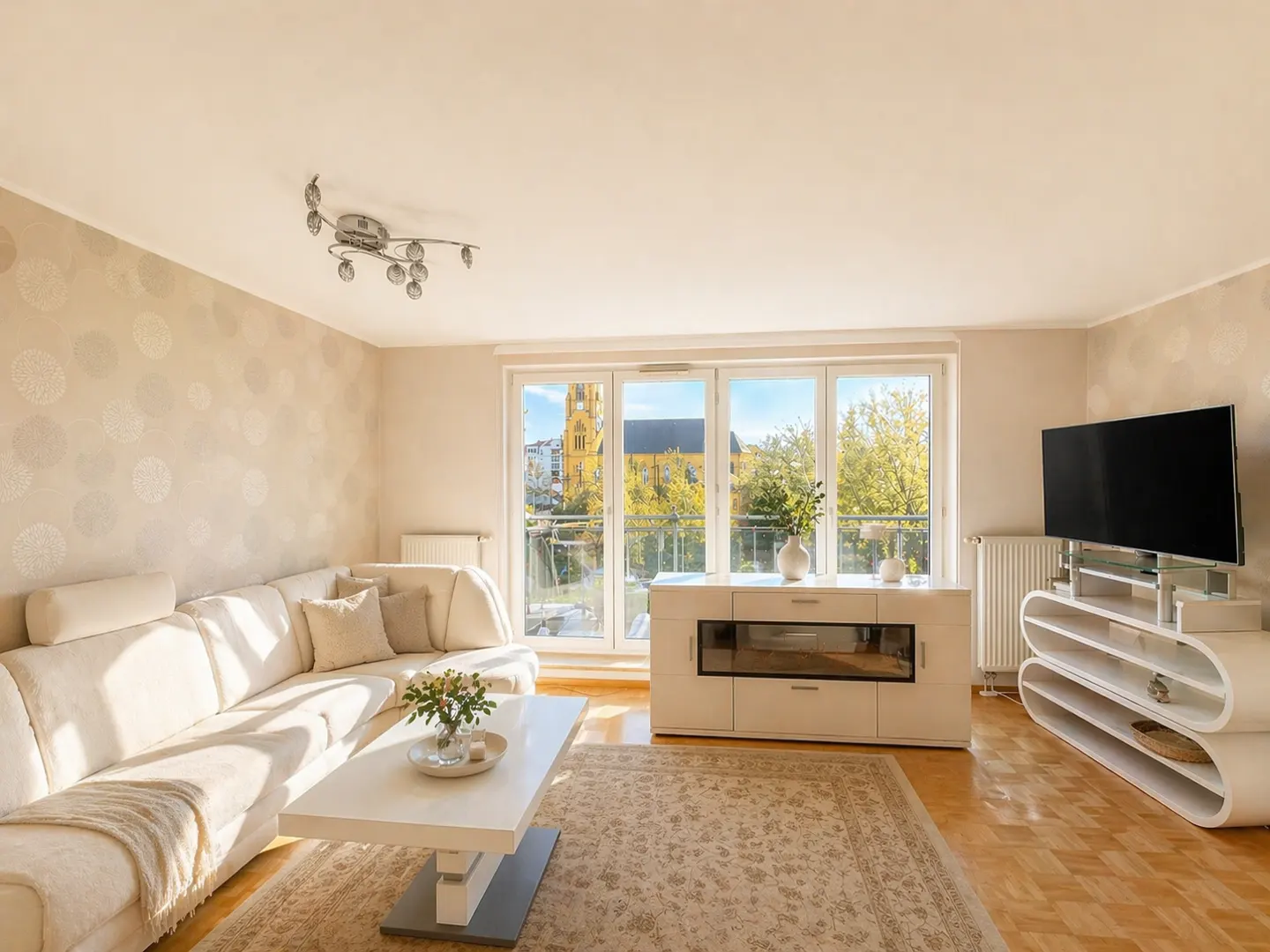 Bright living room with a white sectional sofa, coffee table, modern TV stand, and a view of a yellow church through windows.