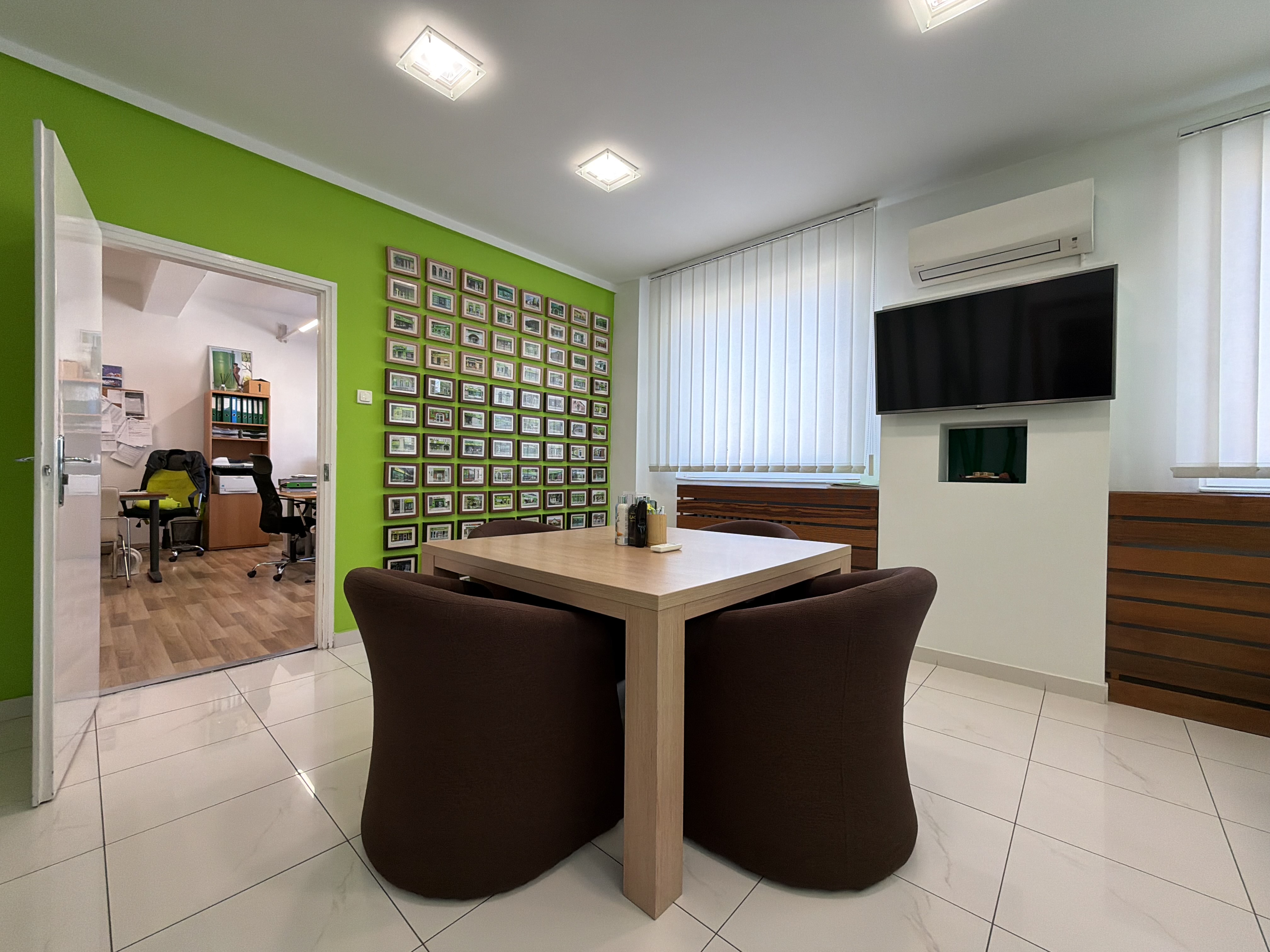 Office interior with a square table surrounded by brown chairs, a green wall with framed pictures, and a TV above a decorative fireplace.