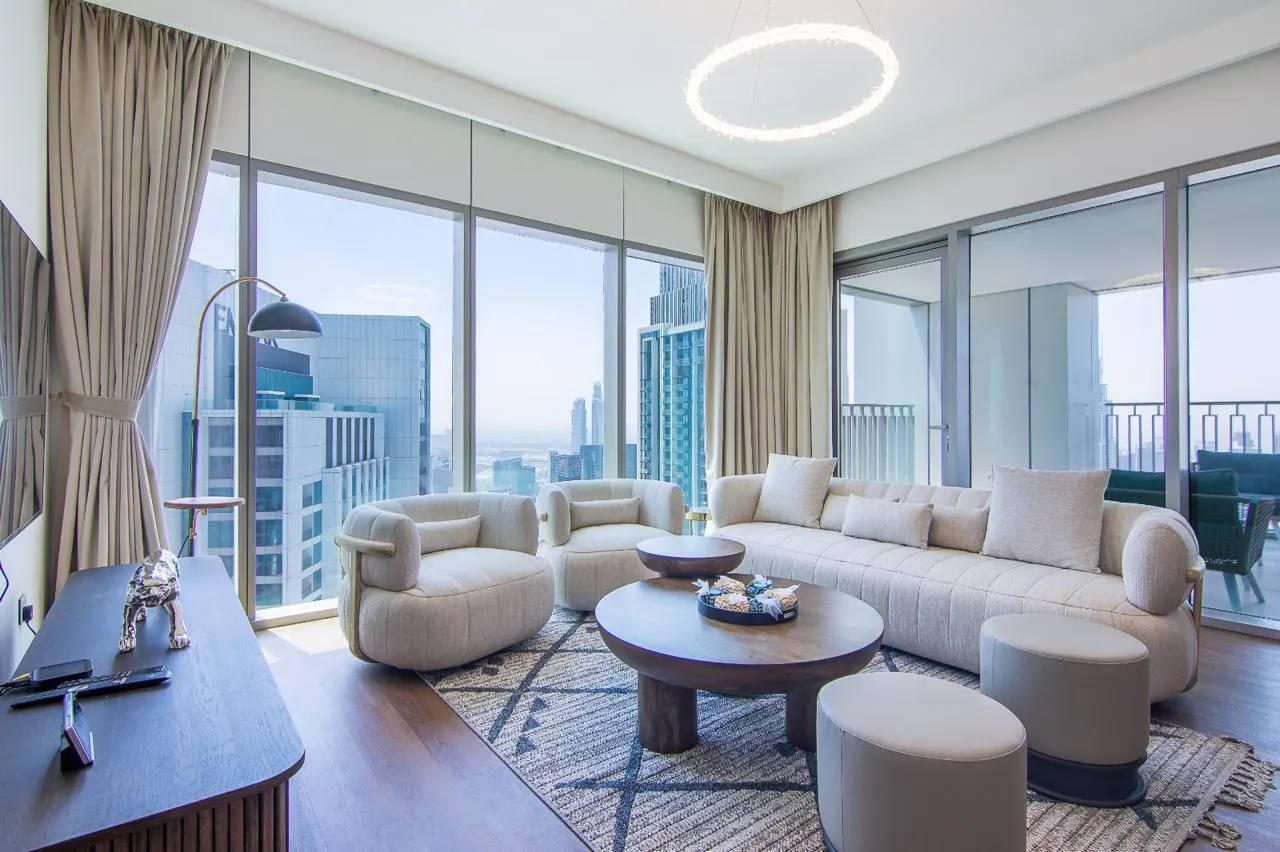 Bright living room with beige sofa, chairs, and rug. Large windows show a city view. A round light fixture hangs from the ceiling.